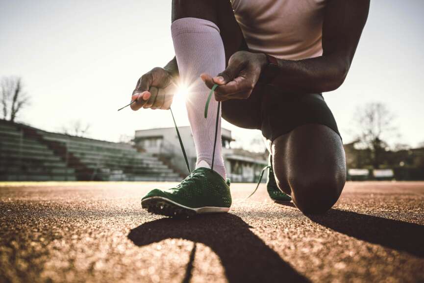 Ein Sportler:innen kniet auf einer Laufbahn, w&auml;hrend er vor dem Training im Sonnenlicht gr&uuml;ne Laufschuhe bindet.