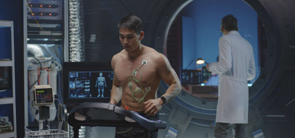 Ein Mann l&auml;uft auf einem Laufband w&auml;hrend eines Leistungstests im Labor und tr&auml;gt dabei &Uuml;berwachungssensoren, w&auml;hrend ein Forscher im Hintergrund beobachtet.