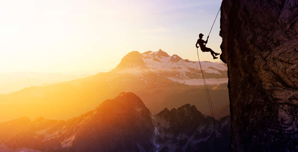 Climbing Trends 2026: What Is Really Changing in Climbing Gear — and What You Will Notice at the Crag Right Away Gear 1 Silhouette of a climber rappelling down a steep rock face at sunset, with glowing alpine peaks and snow-covered mountains in the background.