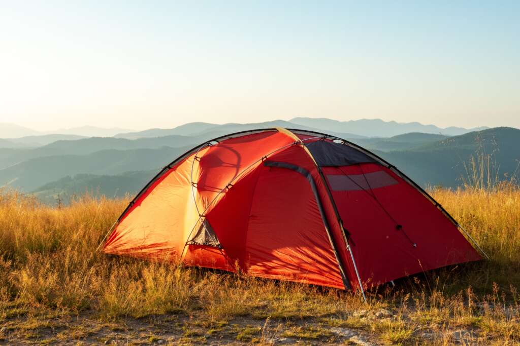 A red camping tent stands in a grassy mountain landscape during golden hour, with rolling hills and distant blue mountain ranges under a clear sky.