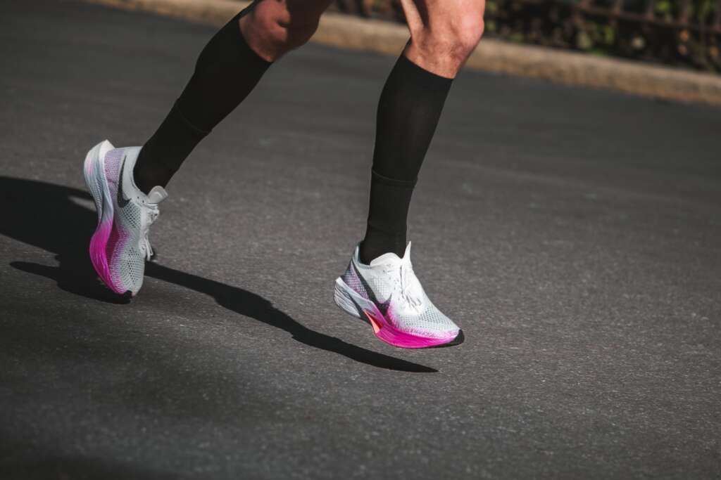 A side-view close-up of a runner's legs mid-stride on asphalt, wearing white and pink Nike Air Zoom Alphafly performance running shoes and black compression socks.