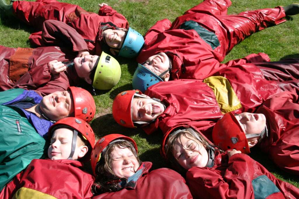 A group of young people in helmets and waterproof clothing lying together on the grass after an outdoor activity.