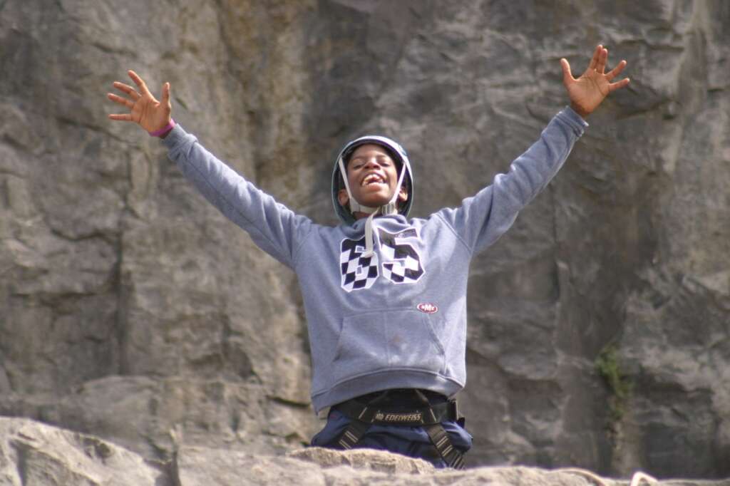 Young person wearing a climbing helmet and harness with arms raised in front of a rock face.